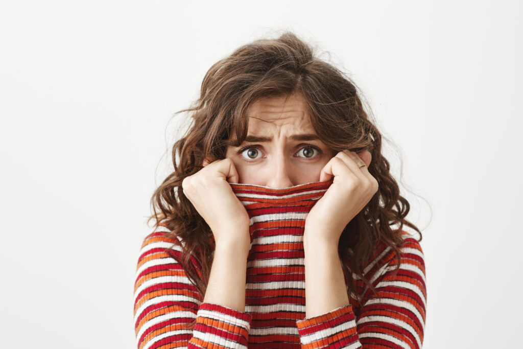 Retrato de una linda joven estudiante escondida en un suéter, mirando a través del cuello con expresión de miedo, frunciendo el ceño y queriendo llorar mientras está de pie sobre un fondo gris.
